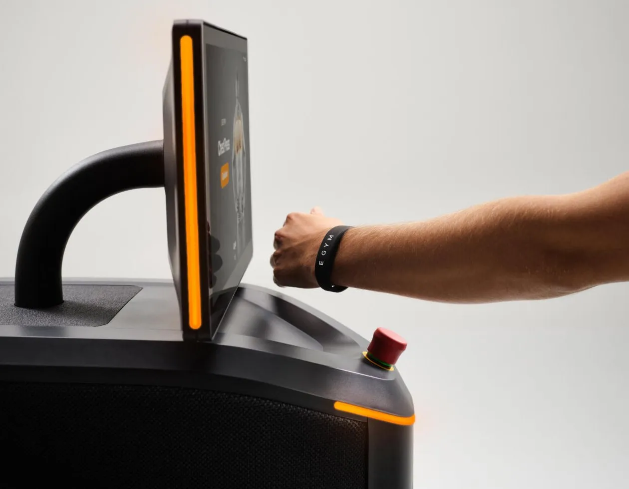 A hand wearing an EGYM wristband taps in at a strength machine's screen, which is edged with an orange light.